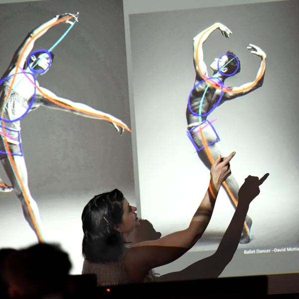 A lecturer points to an image of two ballet dancers in poses on the whiteboard